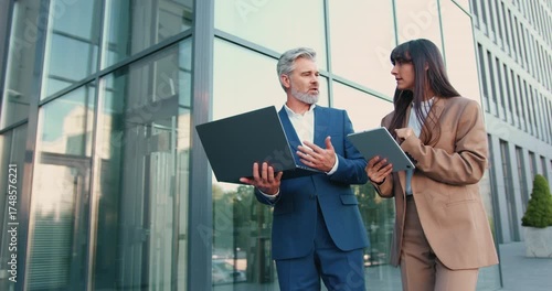 Confident businessman with laptop and elegant businesswoman with tablet walking together down the street toward a modern office building, discussing work and collaborating on a project