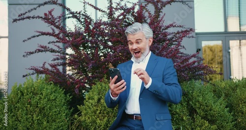 Joyful adult man looking at smartphone with surprised expression, smiling widely and raising eyebrows, standing outdoors.