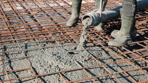 Concrete being poured over steel reinforcement mesh. Construction workers in boots pouring wet concrete onto a rebar grid, preparing the foundation slab for a building structure.