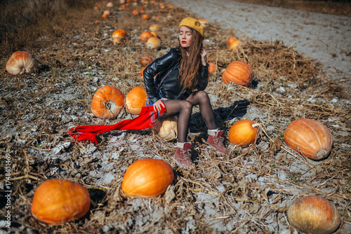 Beautiful young woman, 25 years old, in a black leather jacket and denim skirt, wearing a yellow hat and red scarf, posing stylishly on a pumpkin field in autumn light.