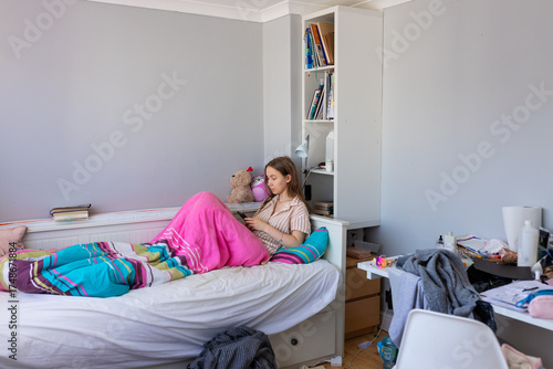 A teenage girl in pajamas lies in bed with her mobile phone in a messy room