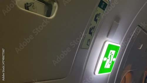Green illuminated emergency exit sign near the entrance door of aircraft, running man with two arrows pointing in both directions, rules and signs on airplane board for safety flight
