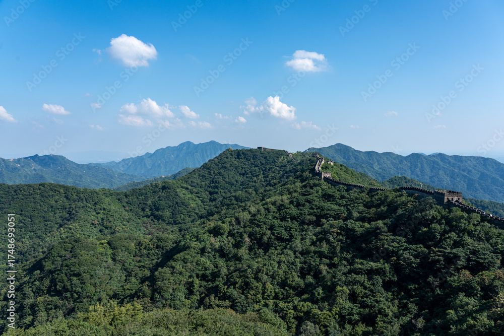 Naklejka premium A wide vista shows the Great Wall of China following the ridge line of a lush, forested mountain range. The impressive structure contrasts with the vast natural landscape under a blue sky 
