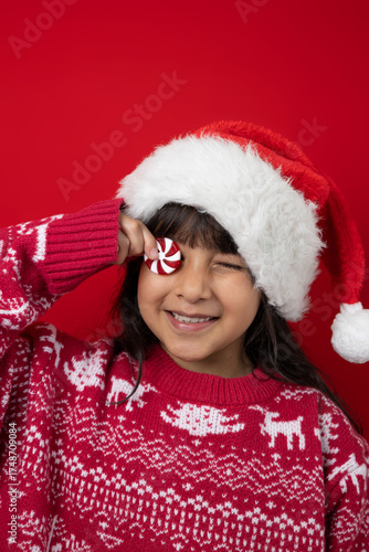 Girl wearing a Christmas hat and sweater playfully covers one eye with a peppermint candy and laughs on a red background. Tender and festive image that conveys joy and Christmas spirit