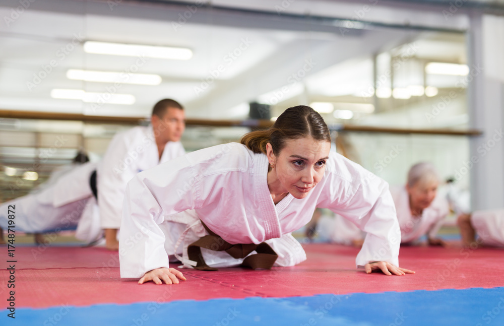 Naklejka premium Focused young woman in white kimono doing pushups during warm up before group martial arts training in gym..