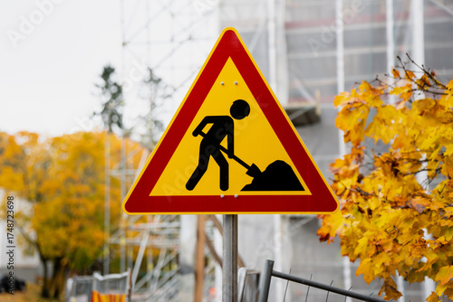 Triangular construction warning sign placed near building renovation site with scaffolding and autumn trees, symbolizing roadwork safety, industrial maintenance and urban infrastructure management