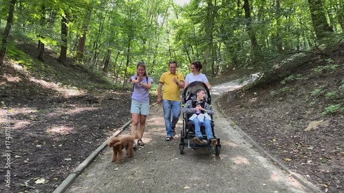 Family with a boy with a central nervous system disorder walking in the park. Family with a boy with inclusion. A boy with a cerebral palsy in a wheelchair walks with his family in the park
