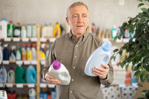 Mature man chooses detergents and liquid laundry gel. Pensioner stands in a store and buys an all-purpose cleaner for surfaces and home