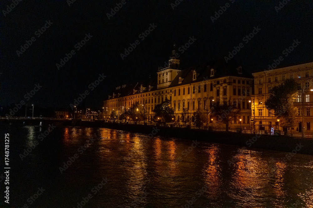 Fototapeta premium Illuminated historic baroque building with clock tower on riverside embankment at night with golden reflections in dark water. Concept of urban nightscape, architectural heritage, romantic atmosphere.