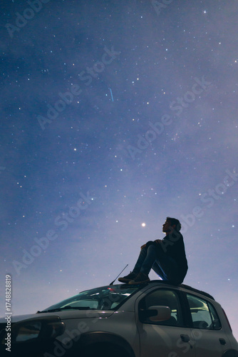 Traveler sitting on a car roof, gazing at the night sky, lost in deep thoughts and spiritual connection beneath the stars.