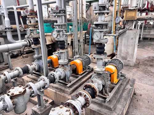 Row of industrial centrifugal water pumps and electric motors connected to complex piping systems, valves, and gauges at an outdoor factory pumping station.