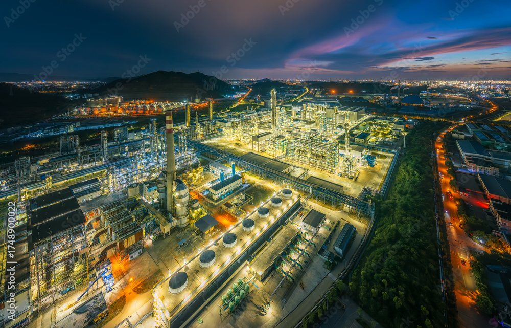 Fototapeta premium Panoramic of a huge industrial complex and petrochemical plant illuminated at night surrounded by green hills and roads.