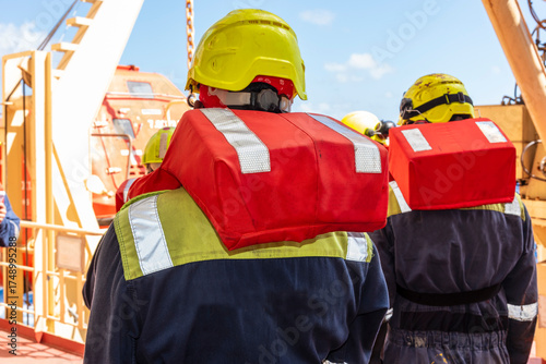 Crew members of merchant ship wear life jackets as they gather for an abandon ship drill, reviewing lifeboat deployment.