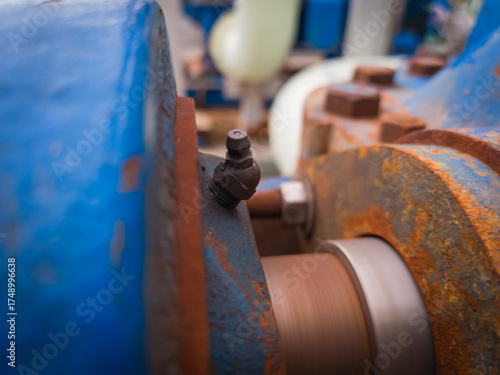 Close-up of a grease fitting (zerk) on rotating industrial machinery. Visible rust, bolts, and motion blur highlight active lubrication in a heavy-duty environment.