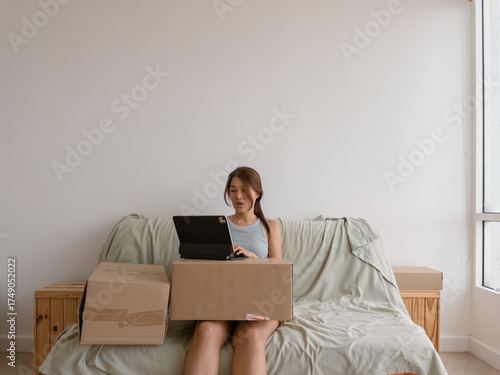Asian woman sitting on bed using laptop with cardboard boxes beside her
