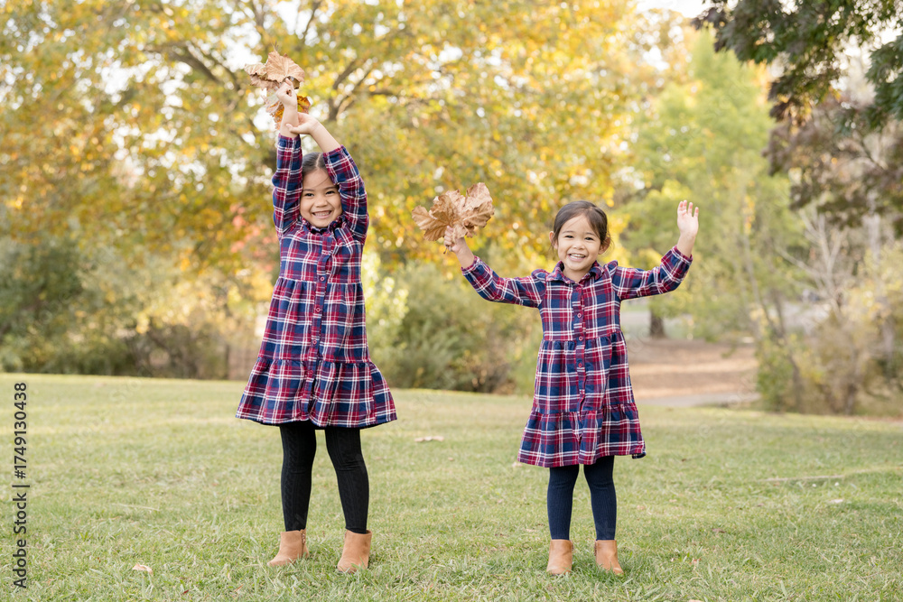 Fototapeta premium Sisters cheerfully raise fall leaves in the park