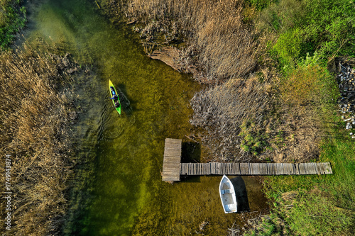 Fototapeta Naklejka Na Ścianę i Meble -  Bird's-eye view of a kayaker passing wooden pier and moored boat on summer lake fringed by reeds in Mazury, Poland