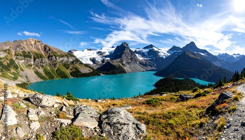 A panoramic vista shows a turquoise lake nestled beneath snow-capped mountains and a cloudy blue sky. Rolling hills of autumn foliage frame the scene