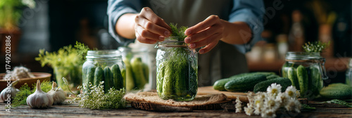 A close-up of hands packing cucumbers into glass jars with dill and garlic on a blurred rustic kitchen background.