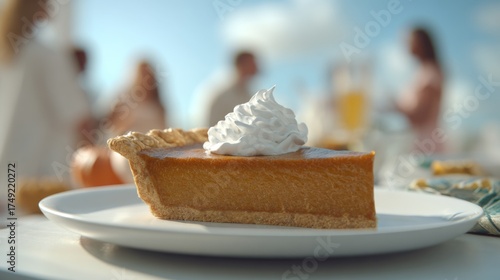 A slice of pumpkin pie topped with whipped cream, set against a blurred background of a festive gathering.