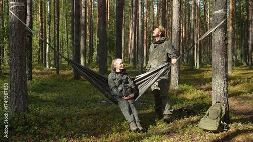Man gently rocks a woman in a hammock between pine trees. They wear outdoor gear and smile. Sunlight filters through the forest. A backpack rests by the tree. Active outdoor recreation