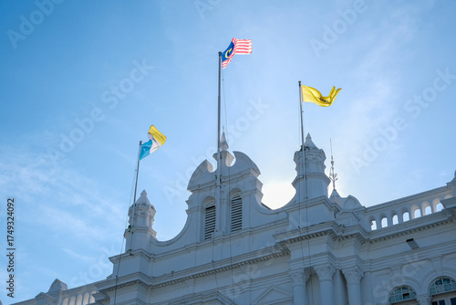 Malaysian national flag and Penang state flag waving above the white colonial architecture style facade of the  George Town Heritage City Municipal City Hall under a bright morning sky.