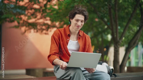 Smiling man sitting outdoors using laptop for video call. Waving hand while talking with friends remotely. Relaxed atmosphere working and socializing in green park area under soft daylight.