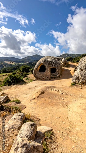 A towering rock formation with openings, situated on a hillside path under a cloudy, vibrant blue sky and rolling hills