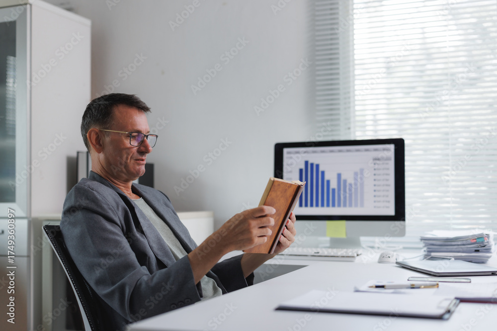 Naklejka premium Mature man reading book while working at office desk