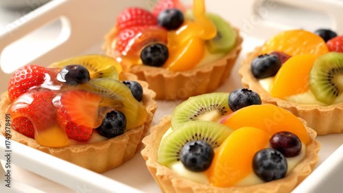 Syrup pouring on delicious fruit tarts with strawberries, blueberries, kiwi, and peaches on plate. Sweet pastry dessert footage.