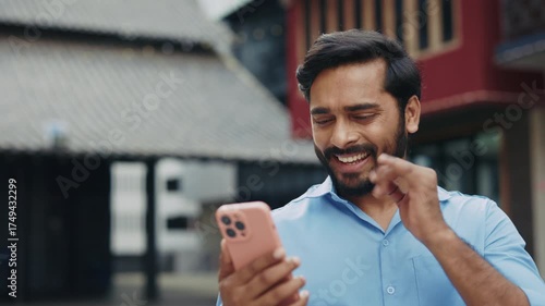 Portrait of happy Indian entrepreneur waves hand Hello and uses sign language on video chat in city. Man with deafness talks on smartphone by gestures standing at urban block