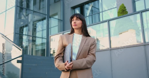 Confident young woman in business suit holding laptop, standing in front of modern office building, smiling softly as she looks into the distance.