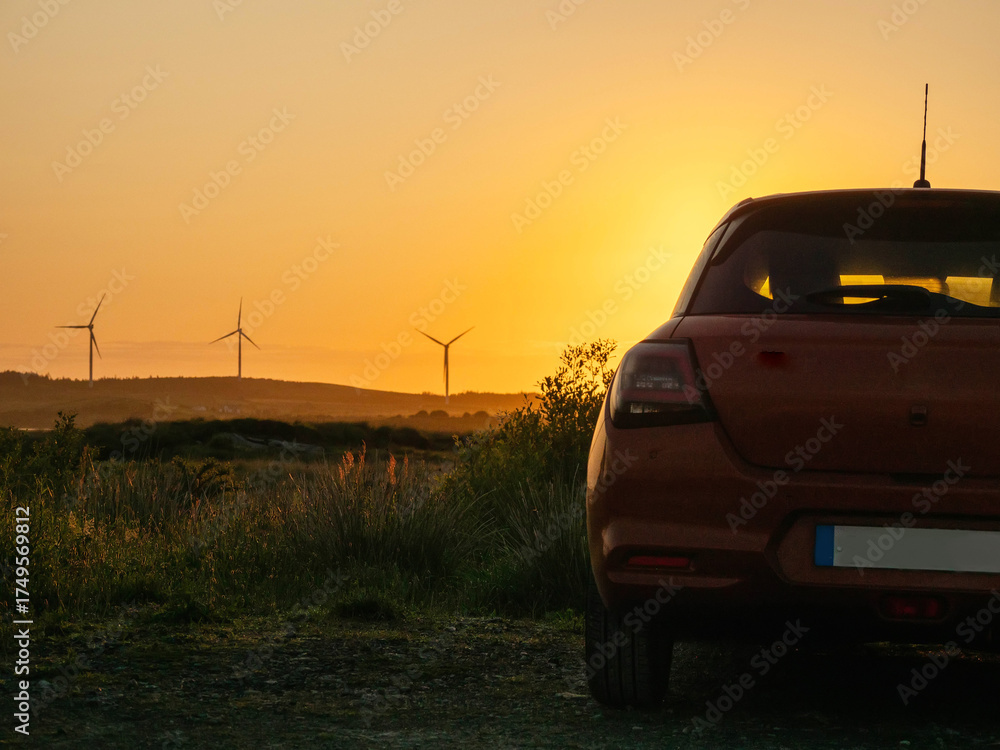 Fototapeta premium A red car is parked in a field with a sunset in the background. The car is facing the sun, and the sky is orange and pink. Wind farm turbines in the background. Sightseeing modern technology concept