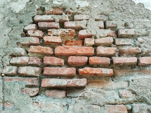 Close-up of a red brick wall in very old and damaged condition, where the cement plaster has peeled off, creating irregular depressions that reveal the arrangement of the red bricks underneath.