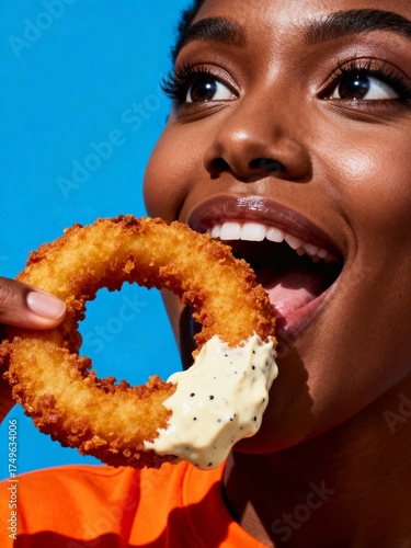 A young woman with dark skin takes a bite of a crispy onion ring dipped in sauce
