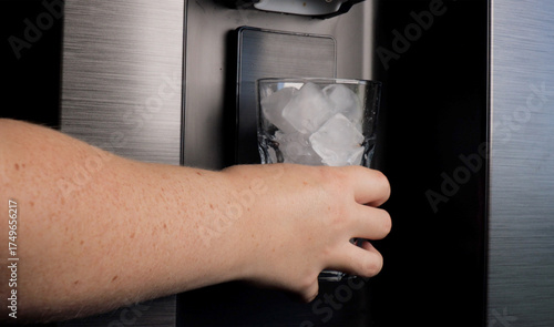 Hand holding a glass with ice cubes under refrigerator dispenser
