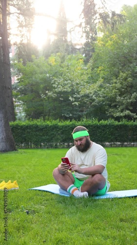 Vertical 4K street video. Fat white man with beard, yellow sport sneakers, green shorts, t-shirt sits on blue mat, watches workout on mobile phone, yoga pose, video, read news, sms in city park. Relax