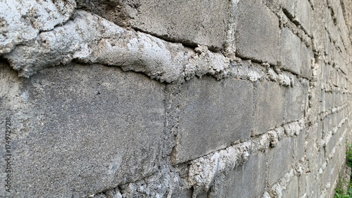 Detailed side-angle view of a weathered exterior wall constructed from rough gray concrete cinder blocks joined by thick, unevenly textured white mortar. Construction concept