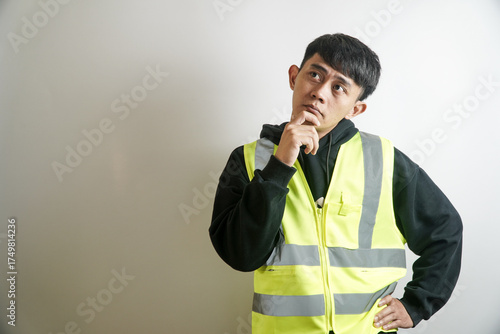 Young Asian industrial worker in a yellow safety vest looking thoughtful and confused, contemplating a solution to a problem