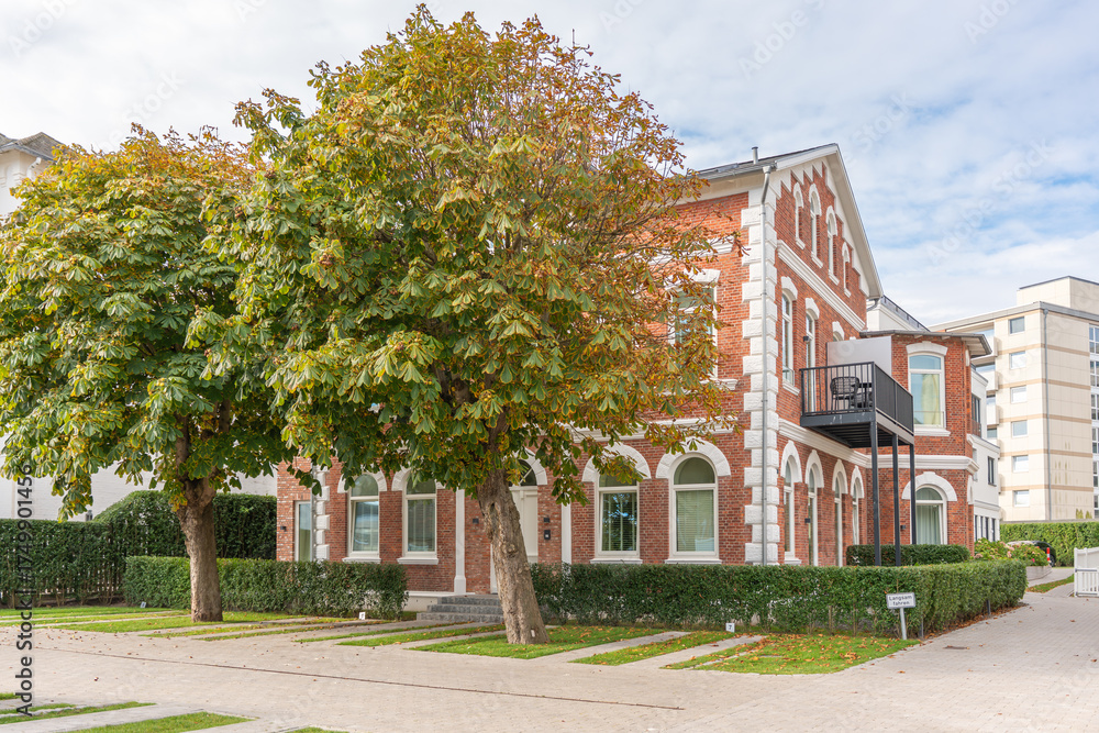 Obraz premium Brick house with trees. A red brick house with white details stands behind large green chestnut trees. The scene feels calm and residential on a sunny day.