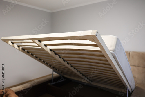  Lift-up bed frame with open storage mechanism in a modern bedroom. Wooden slats and metal supports visible. Practical furniture design and smart home interior concept. High quality photo