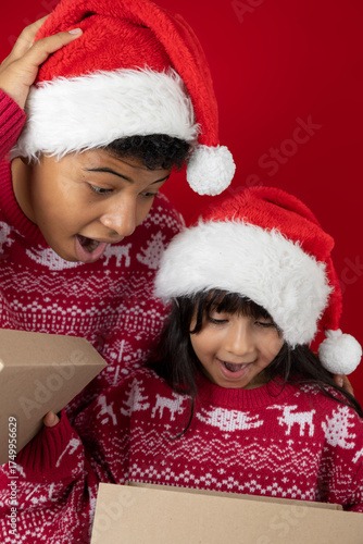 Dark skinned girl and teenage boy wearing Christmas hats and sweaters open a Christmas gift box, surprised with their mouths open on a red background. Ideal image for holiday advertising and campaigns