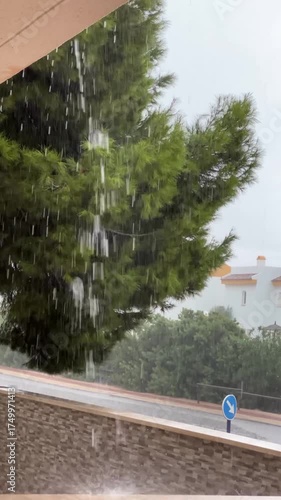Rain pours from the roof, streaking past a dense pine and splashing onto the terrace ledge. A wet road, stone wall, and blue keep‑right sign complete the moody, stormy street view