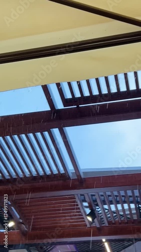 Diagonal view from beneath a cream awning as rain falls in bright streaks. Drops splash off a timber pergola and guttering, with pale sky beyond. Moody, cozy patio moment