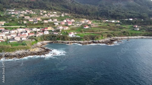 Aerial drone footage flying over Santa María de Oia, capturing a picturesque coastal village with colorful houses, lush green fields, rugged cliffs, and waves crashing on the rocky shoreline  
