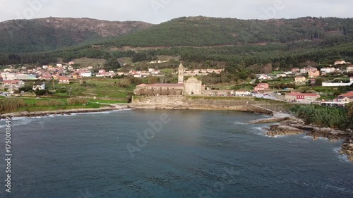 Aerial drone footage approaching the Monastery of Santa María de Oia, highlighting its historic architecture, coastal location, and surrounding green fields and rugged shoreline  
