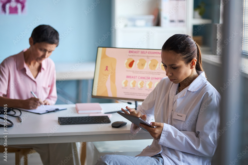 Obraz premium Young adult Caucasian woman doctor using digital tablet while sitting at desk in medical office, middle aged Caucasian woman patient sitting in background with breast cancer awareness chart visible