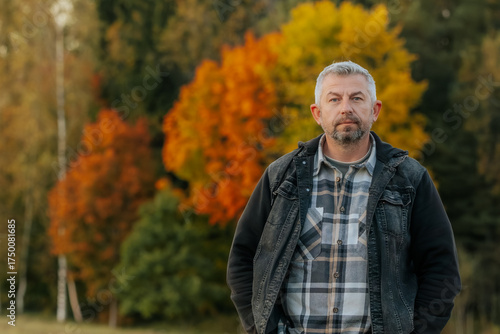 Man standing outdoors in autumn, wearing a plaid shirt and denim jacket, with colorful orange and yellow trees in the blurred background.