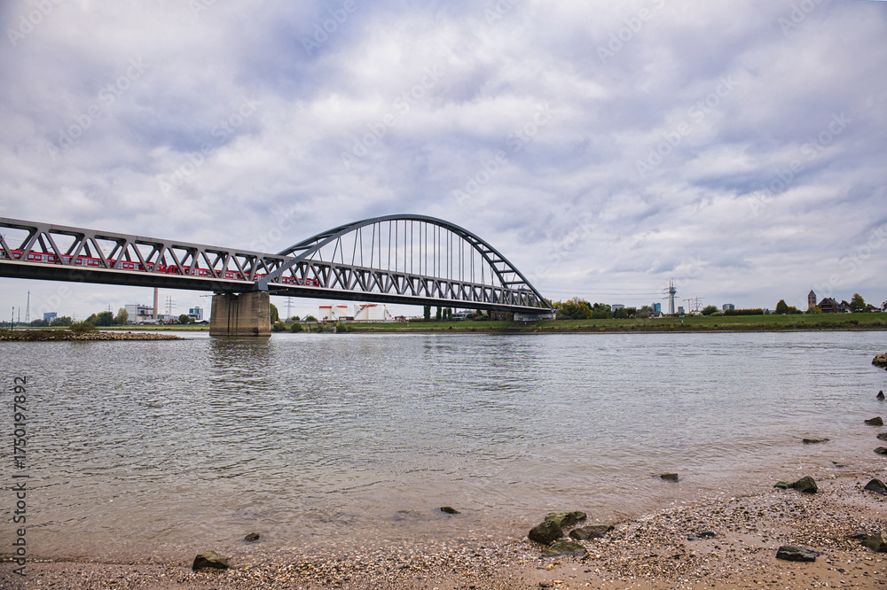 Fototapeta premium Modern steel arch bridge with red commuter train crossing river on cloudy day, transportation and urban infrastructure concept