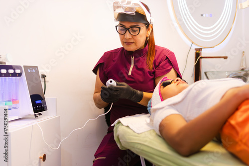A professional aesthetician applies a facial treatment using a device head to a client, with a hydradermabrasion machine visible in the aesthetic treatment room, promoting wellness.